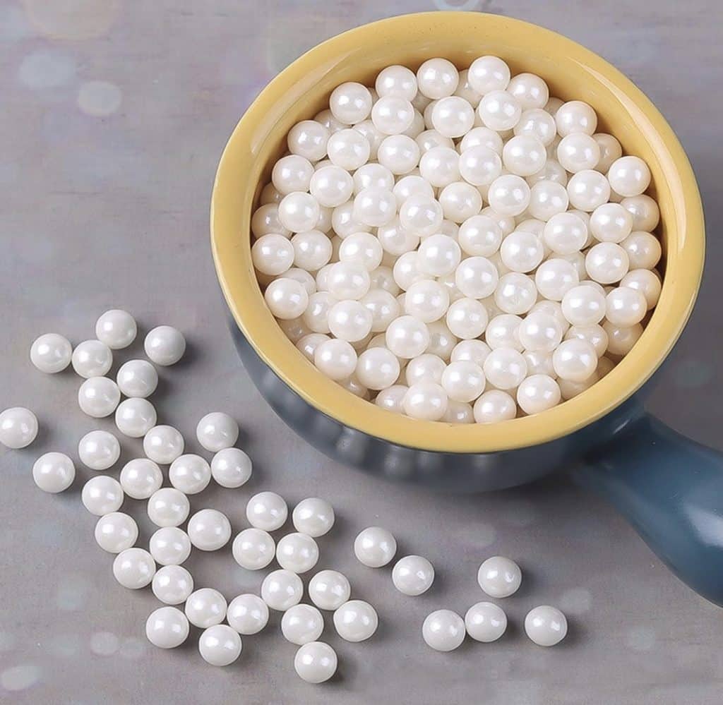 A bowl filled with white pearls sits on a gray surface, resembling the delicate charm of snow globe cookies. Some pearls are scattered outside the bowl, crafting a pleasing arrangement. The bowl is yellow on the inside and blue on the outside.